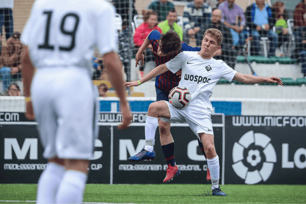 A soccer player in a white kit controlling a soccer ball off his knee during a match.